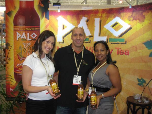 Two women and a man stand together holding bottles of Palo tea in front of a Palo Natural Vitality Tea banner and a large bottle graphic.