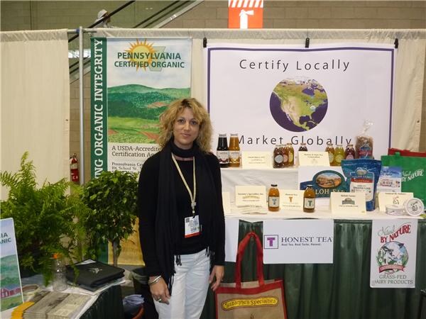 A woman with curly hair and a lanyard stands at a booth with Pennsylvania Certified Organic signs, Honest Tea and Suzanne's Specialties signage, and bottled products on the table.