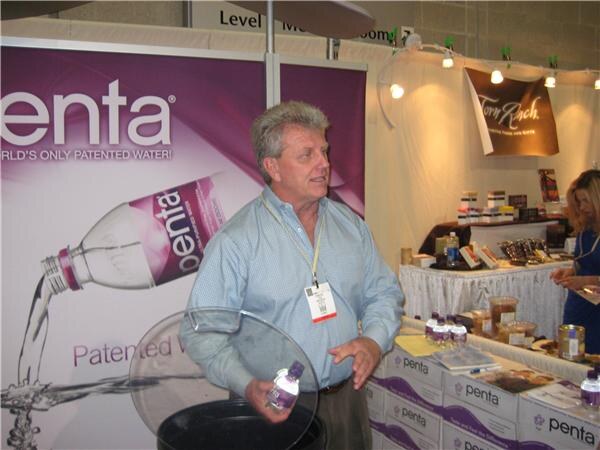 A man in a light blue shirt holds a small Penta water bottle beside a large Penta banner, with cases of Penta and a Torn Ranch display behind him.