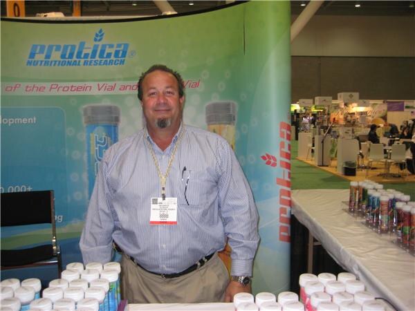 A smiling man in a striped shirt and lanyard stands in front of a Protica Nutritional Research banner, with containers of product on the table in front of him.