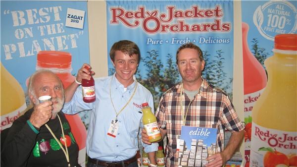 Three men at the Red Jacket Orchards booth hold juice bottles and a copy of edible magazine; the man at left raises a small cup. Banners show Red Jacket juice bottles.