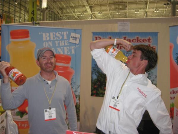 Two men at the Red Jacket Orchards booth playfully hold juice bottles, one raising a bottle overhead and the other tipping a bottle to his mouth, in front of juice banners.