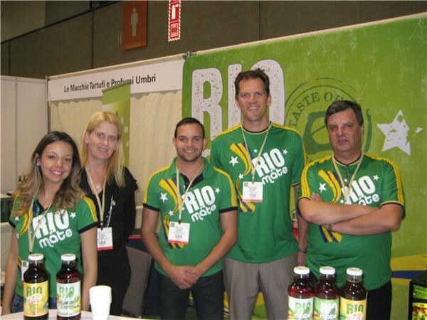 Five people in green Rio Mate shirts and lanyards stand together in front of a green Rio Mate banner, with bottles of Rio Mate drink on the table in front.