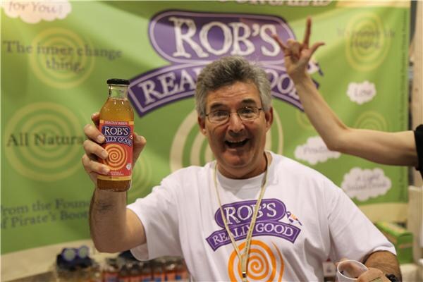 A smiling man with gray hair and glasses holds up a bottle of Rob's Really Good tea in front of a green Rob's Really Good banner. Another person's arm reaches in from the right behind his head.