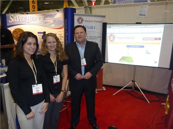 Two women and a man in dark clothing and badges stand on red carpet in front of Silver Biotics and Natural Standard signs and a screen showing the Natural Standard website.