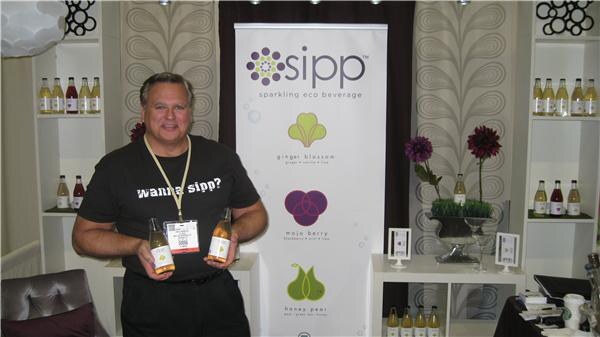 A smiling man in a black "wanna sipp?" T-shirt and a show badge holds two bottles in front of a Sipp sparkling eco beverage banner, with shelves of bottles on both sides.