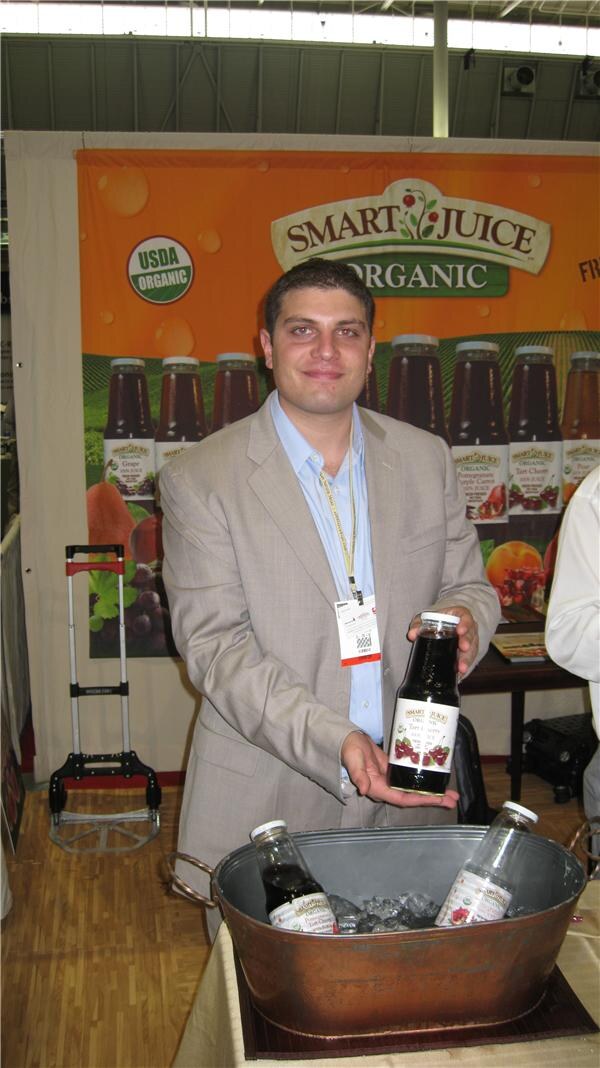 A man in a tan blazer and light blue shirt holds a bottle of dark juice in front of a Smart Juice Organic banner. Two more bottles sit on ice in a copper tub in the foreground.