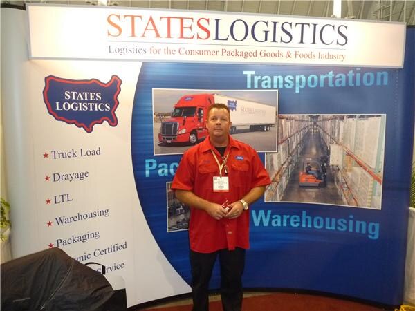 A man in a red short-sleeved shirt and badge stands in front of a States Logistics display listing truck load, drayage, LTL, warehousing and packaging, with images of a truck and a warehouse.