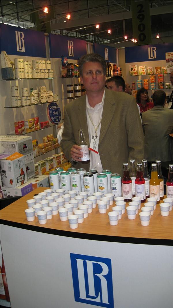 A man in a tan blazer holds a bottle behind a curved counter lined with paper cups, Steaz cans and bottles. Shelves of packaged foods and a large LR logo are visible behind him.