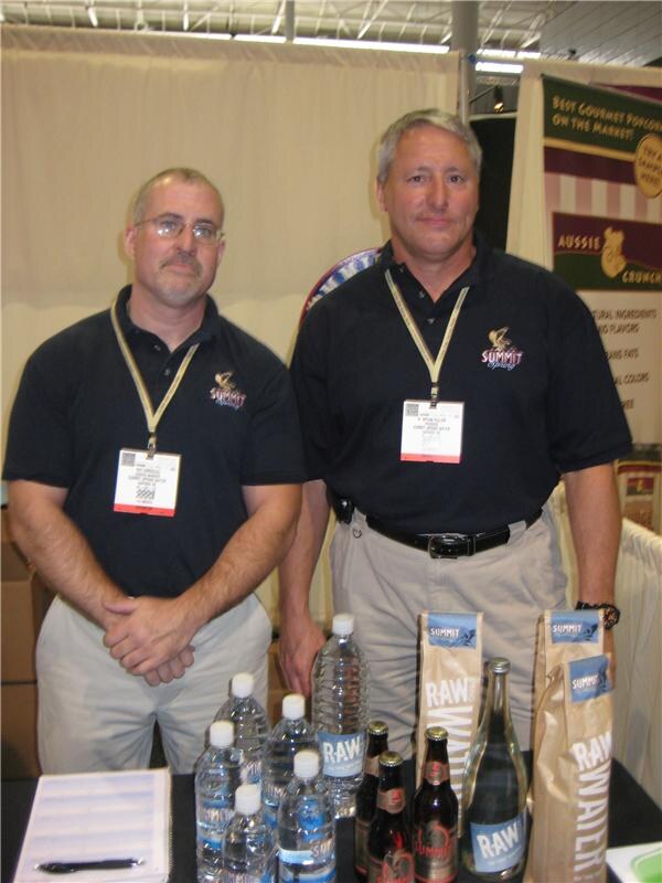 Two men in black Summit Spring polo shirts and badges stand behind a table with bottled water, Raw Water bags and bottles. An Aussie Crunch sign is visible behind them.