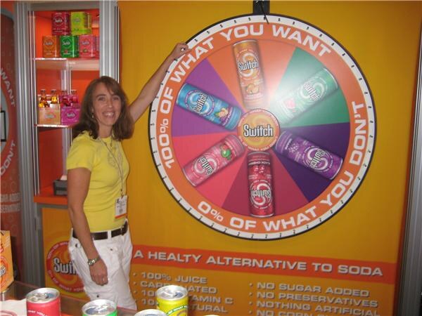 A smiling woman in a yellow shirt reaches up to a large prize wheel showing Switch juice cans. Text on the display reads "100% of what you want, 0% of what you don't."