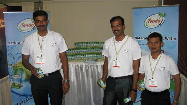 Three men in white Tendo polo shirts and badges stand in front of a table stacked with small cartons, each holding a carton, between two Tendo natural coconut water banners.