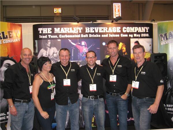 Five men and one woman in black Viva shirts and badges pose in front of The Marley Beverage Company booth backdrop, which lists iced teas, carbonated soft drinks and juices.