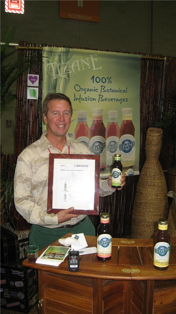 A smiling man in a patterned shirt holds a framed certificate and a Tizane bottle at the Tizane booth, with a Tizane organic botanical infusion banner behind him and bottles on a wooden counter.
