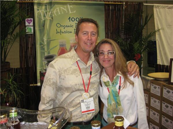 A smiling man and a woman with glasses stand close together, both wearing lanyards, in front of a Tizane banner reading 100% organic infusion beverages, with bottles on the counter below.