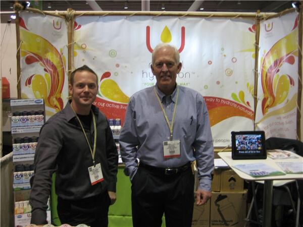 Two men wearing lanyards stand in front of a U Natural Hydration banner with colorful swirls, with product displays at left and a tablet and brochures on a table at right.