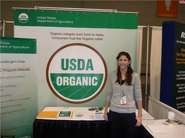 A smiling woman with long dark hair and a badge stands beside a table in front of a large USDA Organic banner from the United States Department of Agriculture.