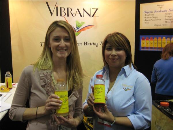Two smiling women each hold a bottle of Vibranz kombucha in front of a Vibranz banner, with a sign listing organic kombucha flavors and a row of bottles behind them.