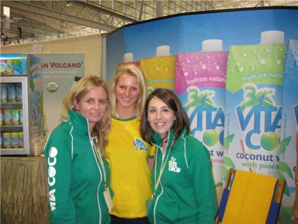 Three smiling women stand together at the Vita Coco booth, two in green Vita Coco jackets and one in a yellow shirt, in front of large Vita Coco coconut water cartons.