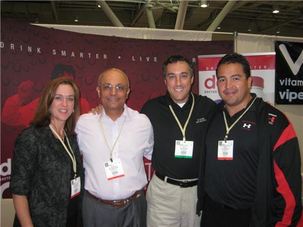 A woman and three men stand close together smiling, wearing lanyards and badges, in front of a dark red banner and a Vitamin Vipers sign.