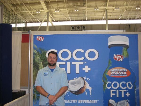 A bearded man in a light blue shirt stands with hands clasped in front of a blue VPX Sports Coco Fit+ banner showing a coconut and a Mangosteen Mania bottle.