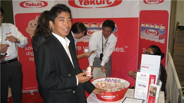 A man in a dark suit holds a small Yakult bottle beside a red bowl of Yakult bottles, with other people and red Yakult banners and brochures behind him.