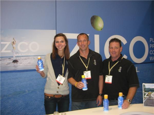 A woman and two men wearing lanyards stand at a counter in front of a large blue Zico banner, holding or standing near bottles of Zico coconut water.