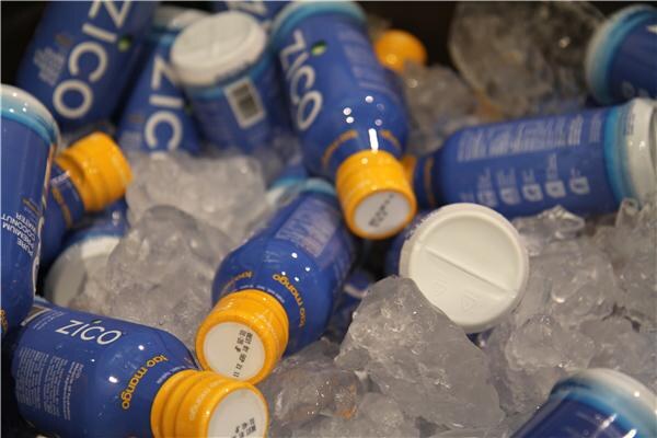 Blue Zico bottles with yellow caps lie on crushed ice, with one bottle labeled mango and a loose white cap among the ice.