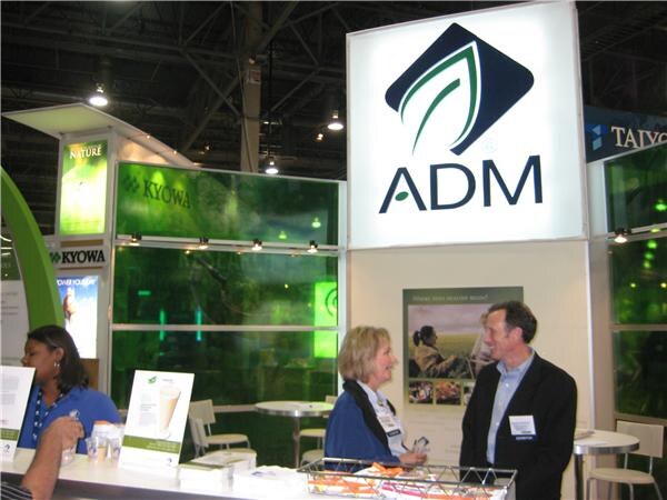 A woman and a man in conversation at a counter beneath a large lit ADM sign. Kyowa signs and green glass panels are behind them, and another woman stands at the left.