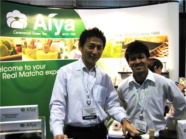 Two smiling men in white shirts with exhibitor badges stand in front of a green Aiya Ceremonial Green Tea banner reading Pure Matcha since 1888, with product literature in the foreground.