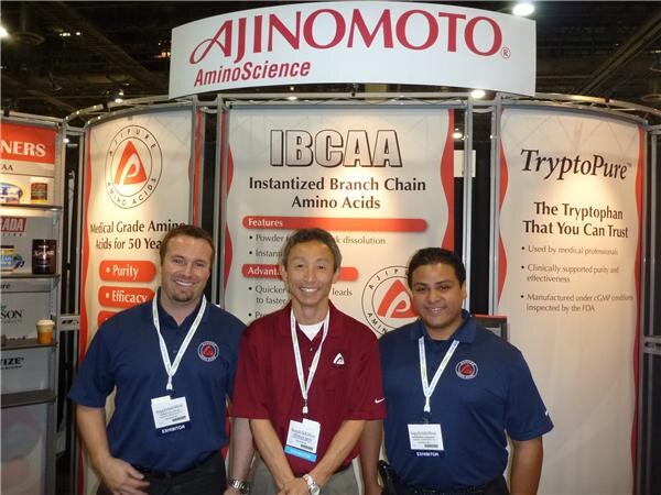 Three men in polo shirts and exhibitor badges stand in front of Ajinomoto AminoScience banners for IBCAA instantized branch chain amino acids and TryptoPure tryptophan.
