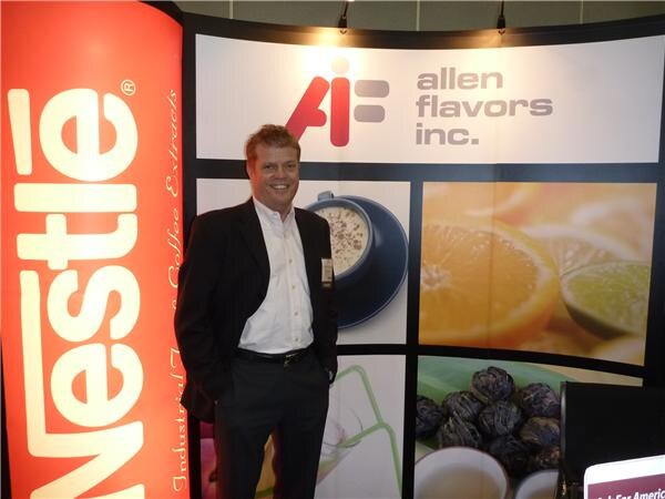 A smiling man in a dark jacket and white shirt stands in front of an Allen Flavors Inc. display showing a coffee cup and citrus slices, beside a tall red Nestle banner.