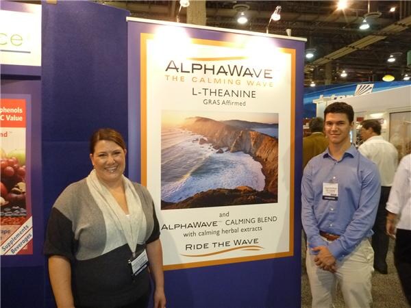 A smiling woman and a young man in a blue shirt stand on either side of an AlphaWave L-Theanine poster showing a coastal cliff and ocean, with the text 'Ride the Wave'.