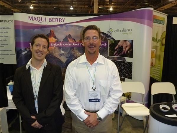 Two men in dark jackets and white shirts stand in front of an Altalena banner with Maqui Berry text and a mountain landscape. One wears an exhibitor badge.