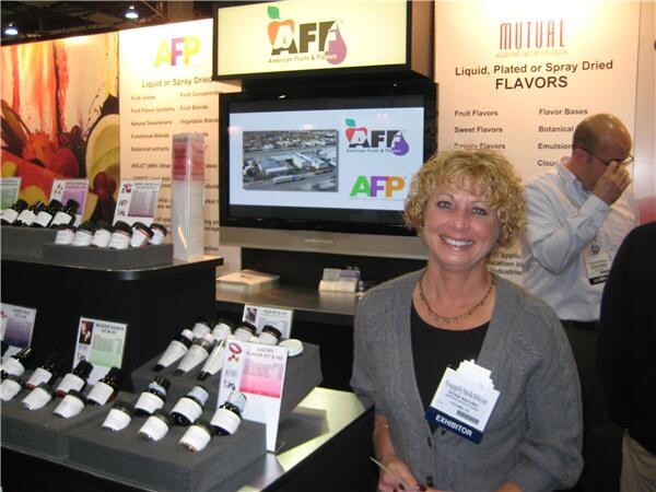 A smiling woman with curly hair and an exhibitor badge stands before a display of flavor sample bottles under American Fruits and Flavors, AFP and Mutual signs. A man stands behind her.