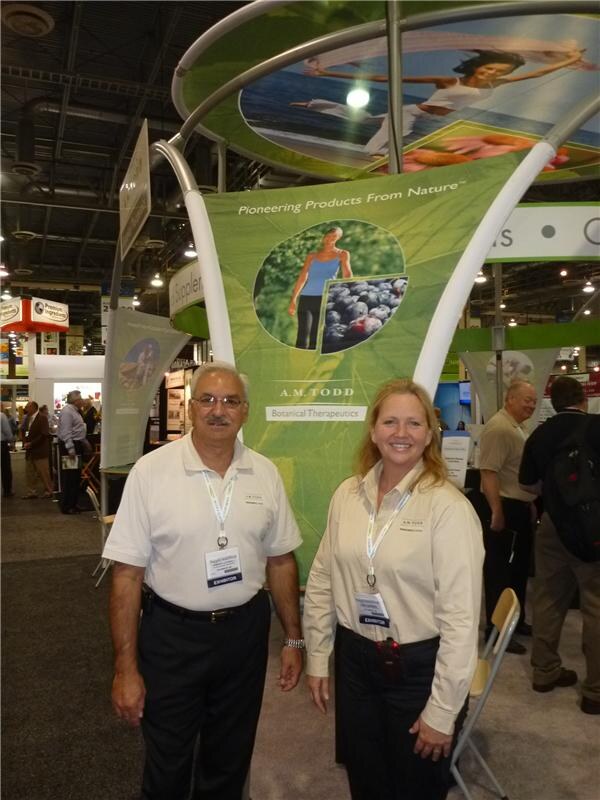 A man in a white polo and a woman in a tan shirt, both wearing exhibitor badges, stand in front of a green A.M. Todd Botanical Therapeutics banner reading Pioneering Products From Nature.
