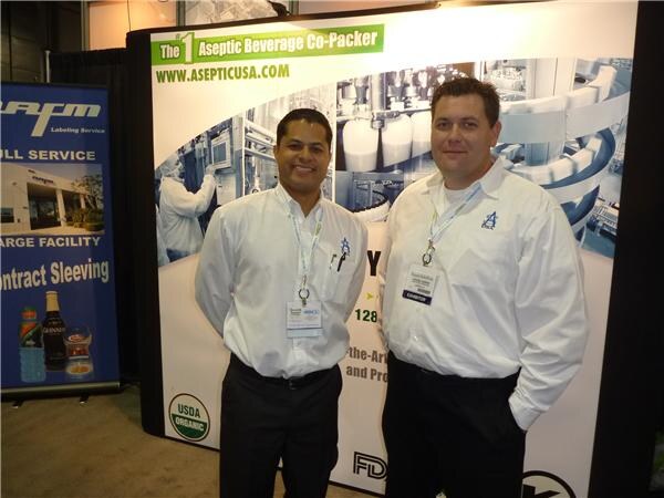 Two men in white shirts with exhibitor badges stand in front of an Aseptic USA banner reading The #1 Aseptic Beverage Co-Packer. An AFM labeling banner is at the left.