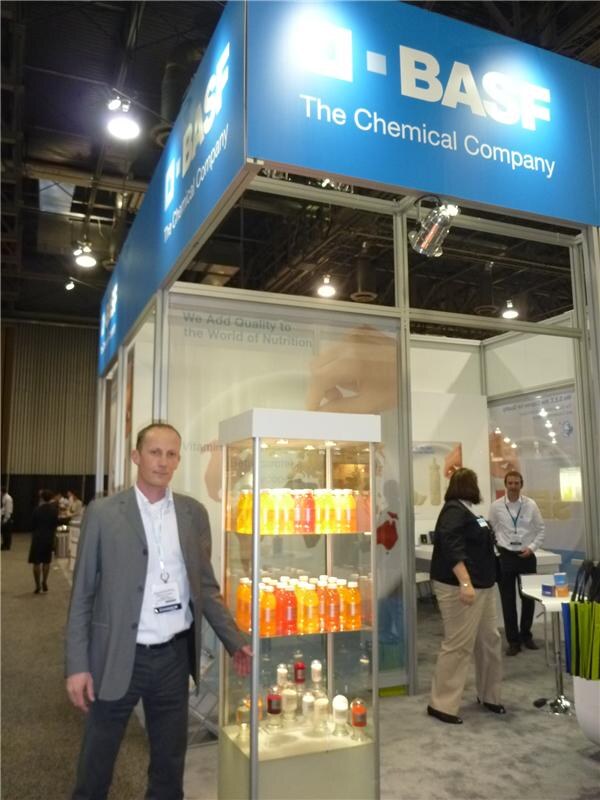 A man in a gray jacket stands beside a lit display case of orange and red beverage bottles in a blue BASF booth marked The Chemical Company. Other people stand in the background.