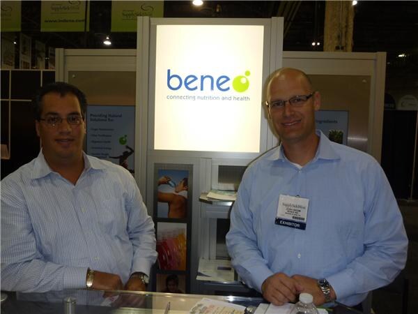 Two men in light blue button-up shirts stand smiling at the Beneo booth counter, one wearing glasses and the other an exhibitor badge, in front of an illuminated Beneo sign.