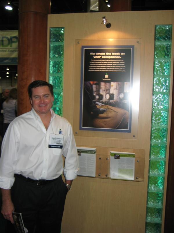 A smiling man in a white shirt and exhibitor badge stands beside a BI Nutraceuticals display with a poster about GMP compliance, green glass block columns and brochures.