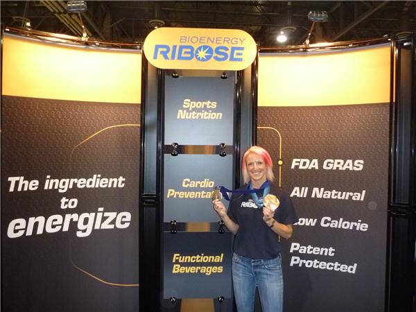 A woman with pink-tinted blond hair holds up two medals on ribbons in front of the Bioenergy Ribose booth, whose panels read The ingredient to energize, FDA GRAS and All Natural.