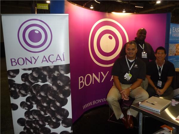 Three men in dark shirts and lanyards sit and stand at the purple Bony Açaí booth, next to a banner showing açaí berries. A laptop rests on the table at right.