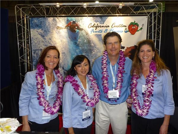 Four people wearing light blue shirts, purple flower leis and exhibitor badges smile in front of a California Custom Fruits & Flavors banner showing a kayaker.