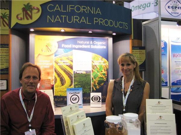 A man in a maroon shirt and a woman in a sleeveless polka-dot top, both with lanyards, stand at the California Natural Products booth with brochures and jars of syrup on the counter.