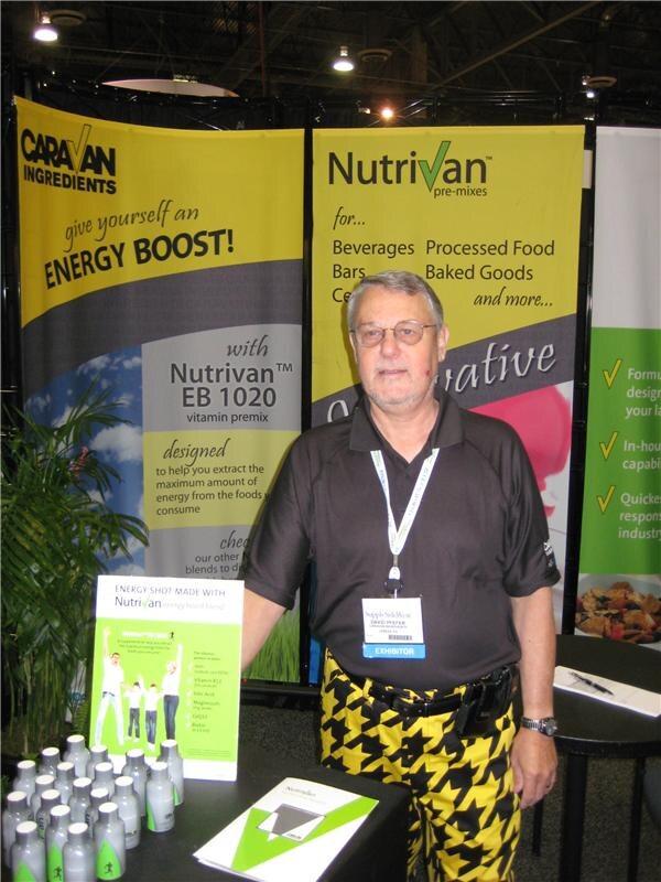 A man with glasses, a black polo and yellow-and-black houndstooth pants stands at a booth with Caravan Ingredients and Nutrivan banners and a display of small energy shot bottles.