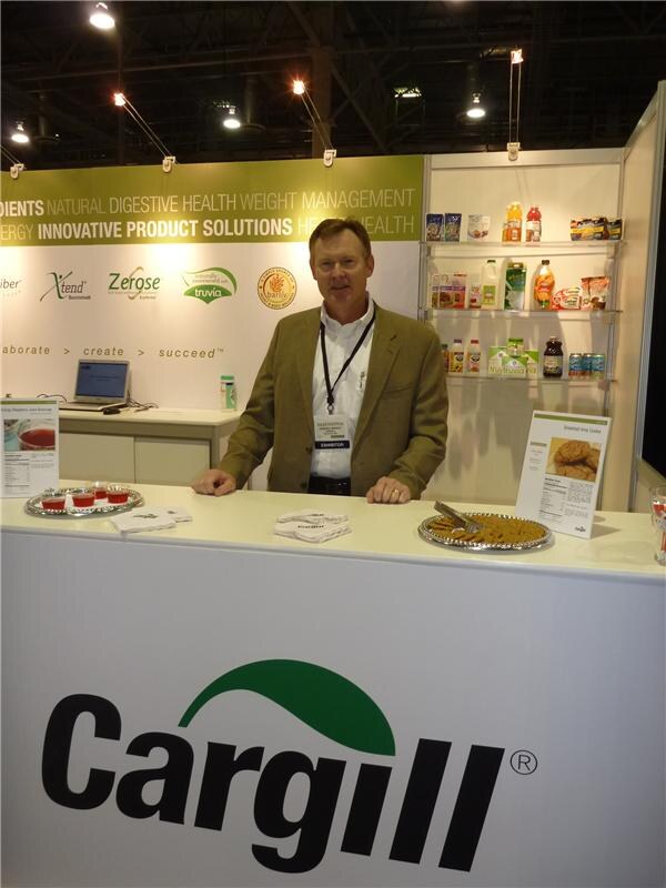 A man in a tan blazer and badge stands behind a white Cargill counter holding trays of samples. Behind him are shelves of products and signs for Truvia, Zerose and Xtend.