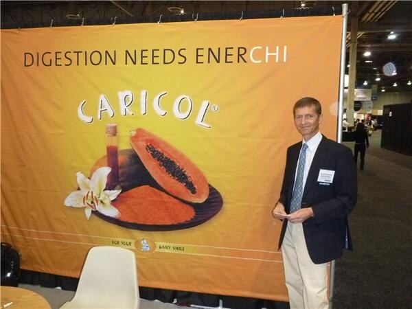 A man in a dark blazer and khaki pants with an exhibitor badge stands beside a large orange Caricol banner showing a halved papaya and a white lily. The banner reads "Digestion needs EnerChi."