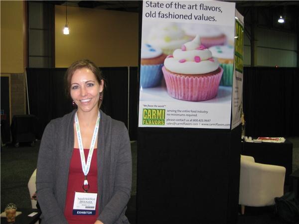 A smiling woman with an exhibitor badge and lanyard stands beside a Carmi Flavors sign showing a pink-frosted cupcake and the text "State of the art flavors, old fashioned values."