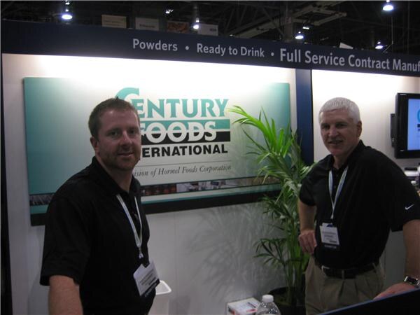Two men in black polo shirts with badges and lanyards stand in front of a Century Foods International sign, with a potted palm and a bottle of water nearby.