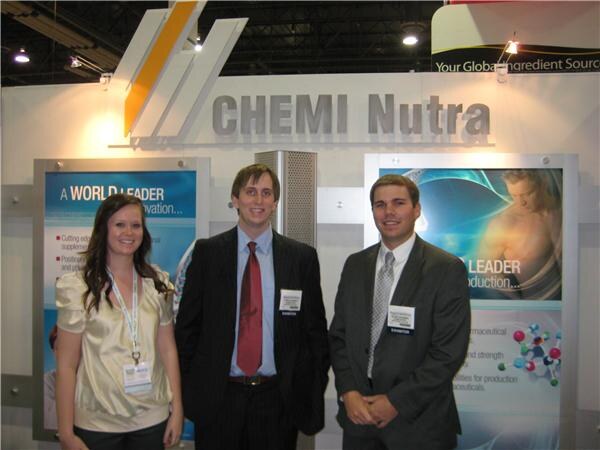 A woman and two men in business attire, all with exhibitor badges, stand in front of a Chemi Nutra sign and display panels with molecule graphics and text about being a world leader.